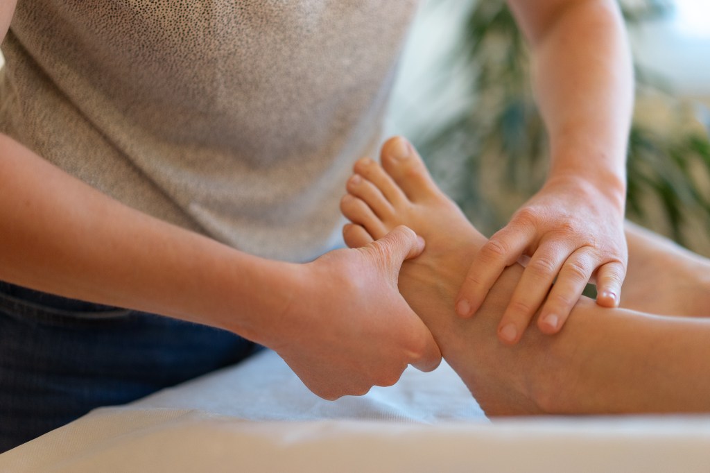 Nahaufnahme der Hände einer Physiotherapeutin, die eine Fussreflexzonenmassage am linken Fuss einer Patientin durchführt.